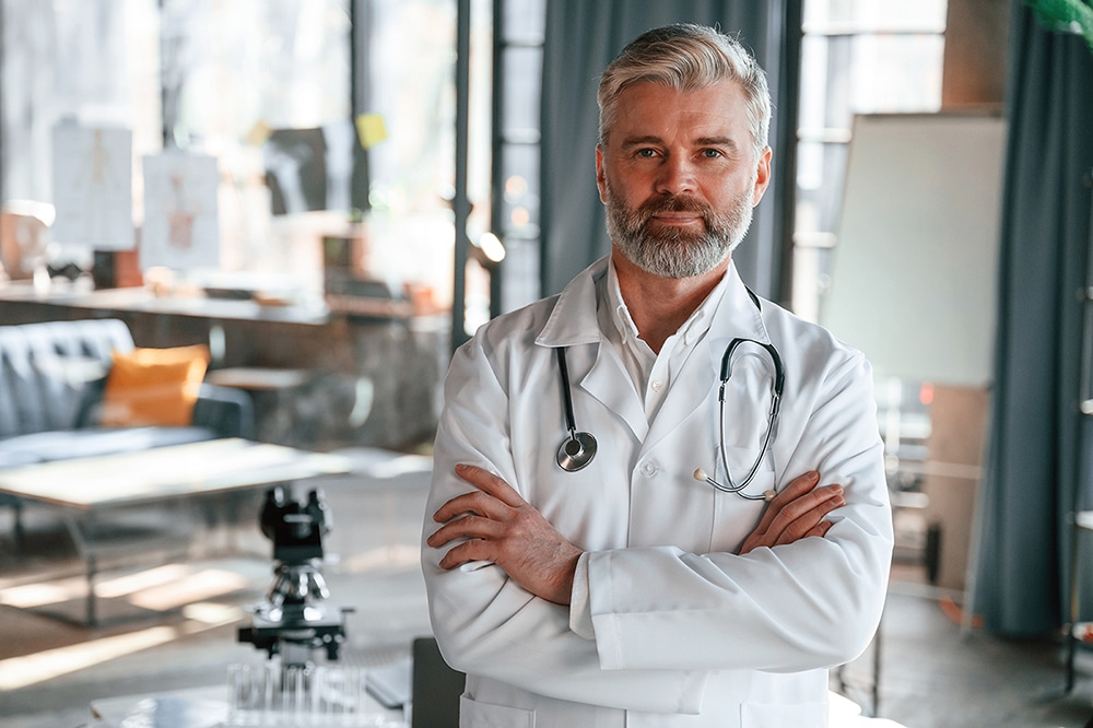 Standing with arms crossed. Senior doctor in white coat is at work indoors content-image
